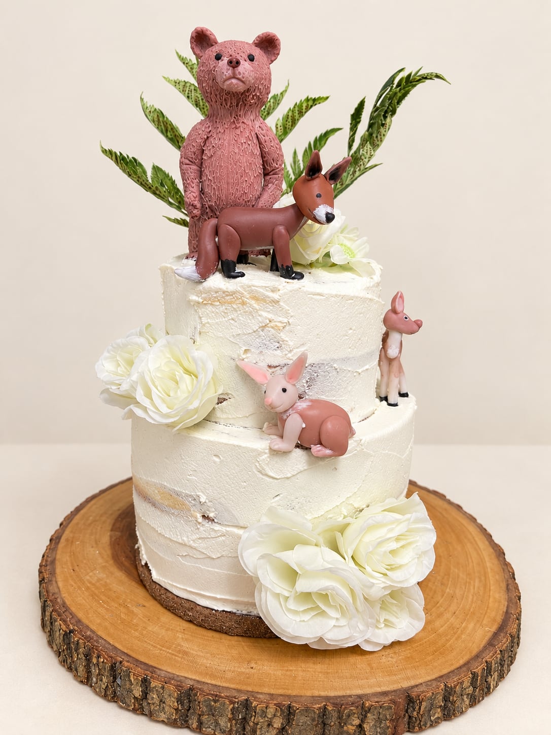 Torta blanca de dos pisos con cuatro figuras de animales, flores blancas y follaje sobre una base de madera.